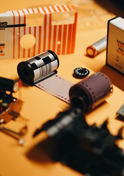 Several film canisters and a strip of exposed film lie on a surface. The items include a mix of film packaging and rolls of both color and black-and-white film. The background is a soft orange color, creating a warm tone for the image.