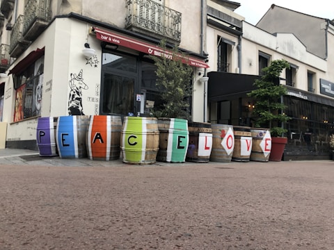 Several barrels are lined up outside a bistro, each painted with a single letter to form the words 'PEACE LOVE'. The barrels feature vibrant colors including purple, blue, orange, green, and red. The building has a rustic charm with intricate iron railings and art painted on the walls.