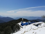 A snow-covered mountainous landscape with a series of blue-roofed buildings, one labeled 'Hotel Munal'. The scene includes expansive views of distant mountain ranges under a clear blue sky with some clouds. A lone person is walking near the buildings.