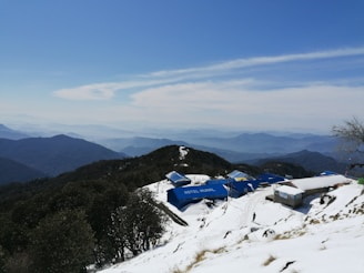 A snow-covered mountainous landscape with a series of blue-roofed buildings, one labeled 'Hotel Munal'. The scene includes expansive views of distant mountain ranges under a clear blue sky with some clouds. A lone person is walking near the buildings.