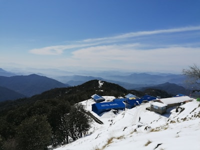 A snow-covered mountainous landscape with a series of blue-roofed buildings, one labeled 'Hotel Munal'. The scene includes expansive views of distant mountain ranges under a clear blue sky with some clouds. A lone person is walking near the buildings.