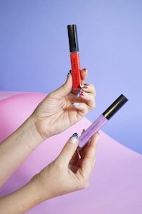 Two hands with colorful nail art holding makeup products, one in red and the other in purple, against a pastel purple and pink background.