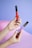 Two hands with colorful nail art holding makeup products, one in red and the other in purple, against a pastel purple and pink background.