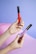 Two hands with colorful nail art holding makeup products, one in red and the other in purple, against a pastel purple and pink background.