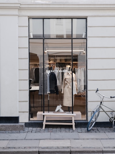 A stylish storefront window displaying well-organized clothing items, including coats and shoes. The window features a clear view into the store, showing various garments hanging neatly. There is a vintage-style bicycle parked against the wall to the right, adding to the urban and chic aesthetic.