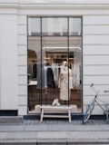A stylish storefront window displaying well-organized clothing items, including coats and shoes. The window features a clear view into the store, showing various garments hanging neatly. There is a vintage-style bicycle parked against the wall to the right, adding to the urban and chic aesthetic.