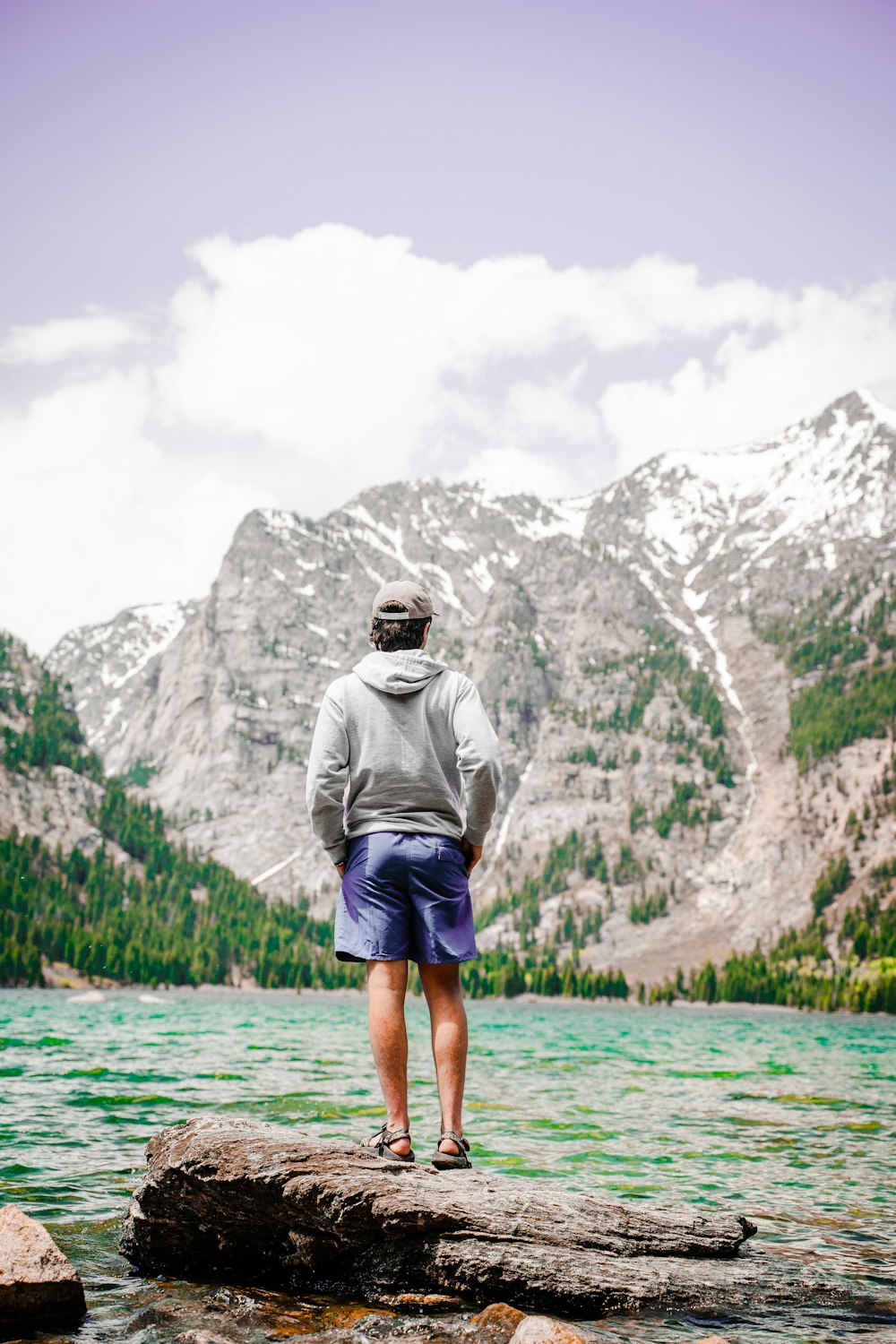 man in grey hoodie standing on green water near grey rocky mountain during daytime