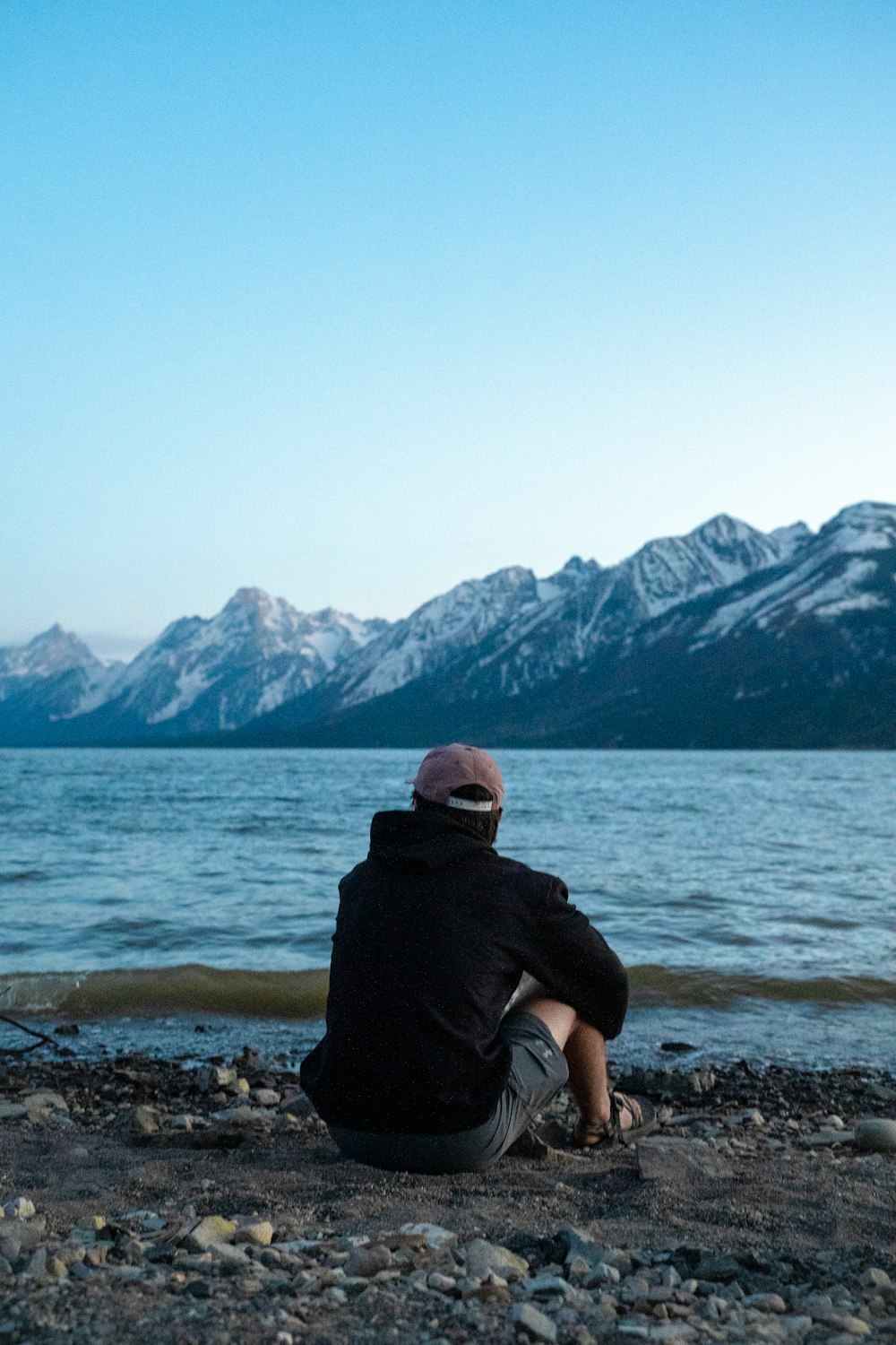 man in black hoodie sitting on rock near body of water and mountains during daytime