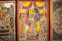 Intricate traditional artwork featuring vividly colored figures in ornate attire, surrounded by detailed patterns and elaborate background designs. The central figure appears in blue, suggesting a depiction of a deity or mythological story, while the surrounding figures are dressed in vibrant saris with elaborate jewelry. The scene is framed by decorative borders, with other artworks partially visible on either side.