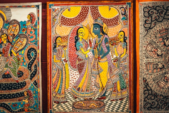 Intricate traditional artwork featuring vividly colored figures in ornate attire, surrounded by detailed patterns and elaborate background designs. The central figure appears in blue, suggesting a depiction of a deity or mythological story, while the surrounding figures are dressed in vibrant saris with elaborate jewelry. The scene is framed by decorative borders, with other artworks partially visible on either side.