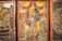 Intricate traditional artwork featuring vividly colored figures in ornate attire, surrounded by detailed patterns and elaborate background designs. The central figure appears in blue, suggesting a depiction of a deity or mythological story, while the surrounding figures are dressed in vibrant saris with elaborate jewelry. The scene is framed by decorative borders, with other artworks partially visible on either side.