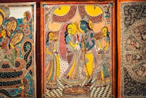 Intricate traditional artwork featuring vividly colored figures in ornate attire, surrounded by detailed patterns and elaborate background designs. The central figure appears in blue, suggesting a depiction of a deity or mythological story, while the surrounding figures are dressed in vibrant saris with elaborate jewelry. The scene is framed by decorative borders, with other artworks partially visible on either side.