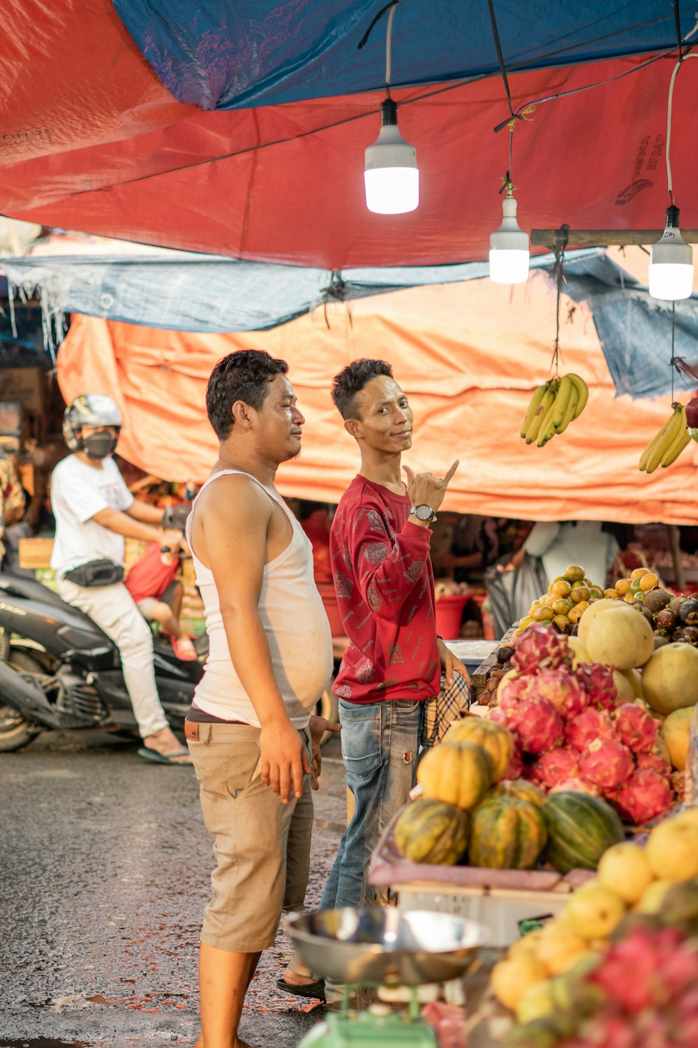 man in red tank top and blue denim jeans standing in front of fruit stand