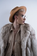 A stylish individual wearing a wide-brimmed tan hat, large round glasses, a fur coat, and a beige shirt. The look is completed with a gold chain necklace and hoop earring, adding an elegant and avant-garde flair.