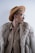 A stylish individual wearing a wide-brimmed tan hat, large round glasses, a fur coat, and a beige shirt. The look is completed with a gold chain necklace and hoop earring, adding an elegant and avant-garde flair.