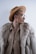 A stylish individual wearing a wide-brimmed tan hat, large round glasses, a fur coat, and a beige shirt. The look is completed with a gold chain necklace and hoop earring, adding an elegant and avant-garde flair.