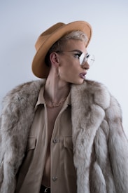 A stylish individual wearing a wide-brimmed tan hat, large round glasses, a fur coat, and a beige shirt. The look is completed with a gold chain necklace and hoop earring, adding an elegant and avant-garde flair.