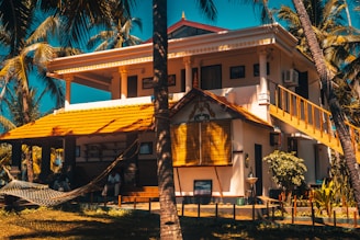 A picturesque two-story house with a vibrant yellow roof set amidst tall palm trees. The house features a spacious balcony and a staircase leading up. A hammock is strung between two palm trees in the lush garden foreground, creating a serene tropical setting.