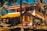 A picturesque two-story house with a vibrant yellow roof set amidst tall palm trees. The house features a spacious balcony and a staircase leading up. A hammock is strung between two palm trees in the lush garden foreground, creating a serene tropical setting.