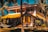 A picturesque two-story house with a vibrant yellow roof set amidst tall palm trees. The house features a spacious balcony and a staircase leading up. A hammock is strung between two palm trees in the lush garden foreground, creating a serene tropical setting.