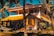 A picturesque two-story house with a vibrant yellow roof set amidst tall palm trees. The house features a spacious balcony and a staircase leading up. A hammock is strung between two palm trees in the lush garden foreground, creating a serene tropical setting.