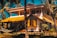 A picturesque two-story house with a vibrant yellow roof set amidst tall palm trees. The house features a spacious balcony and a staircase leading up. A hammock is strung between two palm trees in the lush garden foreground, creating a serene tropical setting.