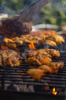 Pieces of seasoned chicken are sizzling on a barbecue grill with visible flames and smoke rising between the grill grates. The scene suggests outdoor cooking, surrounded by a natural green backdrop, creating a warm, inviting atmosphere.