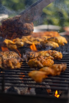 Pieces of seasoned chicken are sizzling on a barbecue grill with visible flames and smoke rising between the grill grates. The scene suggests outdoor cooking, surrounded by a natural green backdrop, creating a warm, inviting atmosphere.