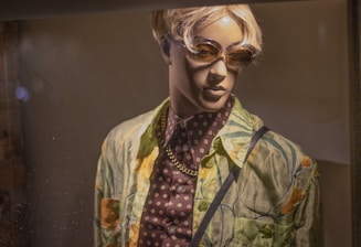A mannequin is dressed in a colorful tropical-themed shirt over a patterned dark shirt, accessorized with a gold chain necklace. It is wearing sunglasses and has short, blonde hair, displayed under warm lighting.