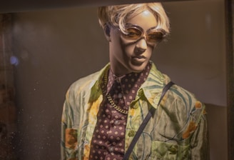 A mannequin is dressed in a colorful tropical-themed shirt over a patterned dark shirt, accessorized with a gold chain necklace. It is wearing sunglasses and has short, blonde hair, displayed under warm lighting.