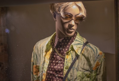 A mannequin is dressed in a colorful tropical-themed shirt over a patterned dark shirt, accessorized with a gold chain necklace. It is wearing sunglasses and has short, blonde hair, displayed under warm lighting.