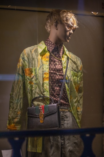 A mannequin dressed in a stylish, patterned outfit stands in a display window. The clothing includes a green and orange floral shirt over a brown polka dot shirt, paired with gray pants. The mannequin wears sunglasses and has a shoulder bag with a chain strap that features green and red stripes along with a golden buckle.