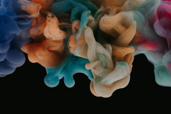 Swirling clouds of colorful smoke or ink create an abstract pattern against a dark background. Various colors, including blue, orange, teal, and hints of red, blend and intertwine to form soft, flowing shapes.