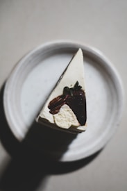 A slice of cheesecake is placed on a white ceramic plate. The cheesecake is topped with a chocolate-dipped strawberry and pecans. The lighting is soft, casting shadows on the plate and emphasizing the textures of the cake and toppings.