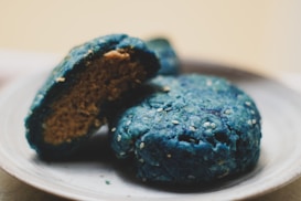 Two round blue cookies are placed on a white plate. One cookie is whole while the other is partially broken to reveal a golden brown interior. Both cookies are speckled with small white seeds on their surface.