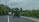 Two vehicles are driving on a paved rural road surrounded by lush greenery and overcast skies. The vehicle in the foreground is carrying a large load of green bananas, partially visible through the open back hatch. Dense vegetation lines both sides of the road.