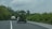 Two vehicles are driving on a paved rural road surrounded by lush greenery and overcast skies. The vehicle in the foreground is carrying a large load of green bananas, partially visible through the open back hatch. Dense vegetation lines both sides of the road.