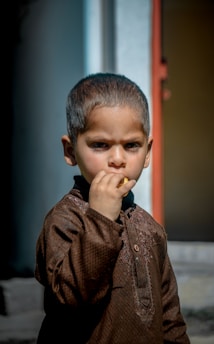 A young child wearing traditional brown clothing is standing outdoors with a serious expression, holding a small object in one hand near the mouth. The background features a blurred doorframe and building structure.