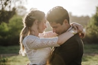 A couple embraces tenderly in a natural setting. The woman, with braided hair, wears a white lace top, and the man wears a dark jacket. Sunlight illuminates their faces, creating a warm glow. The background consists of greenery and soft focus foliage.