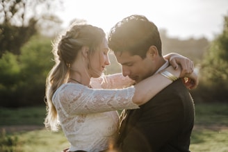 A couple embraces tenderly in a natural setting. The woman, with braided hair, wears a white lace top, and the man wears a dark jacket. Sunlight illuminates their faces, creating a warm glow. The background consists of greenery and soft focus foliage.