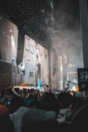 A large crowd is gathered in front of a stage with massive screens displaying images of people and furniture. Confetti is falling from above, adding to the celebratory atmosphere. The audience members are holding up lights and banners, creating an energetic and lively scene.