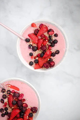 Close-up of a gluten-free, sugar-free pink smoothie bowl topped with fresh berries and chia seeds on a white and light brown background.