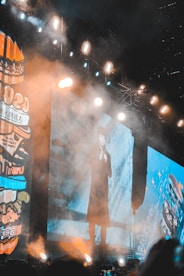 A large stage setup with bright, intense lighting beams across the scene, illuminating a large screen displaying a person in a long coat. Smoke or fog adds a dramatic effect, while vibrant colors are present on the side panels displaying graphics and text.