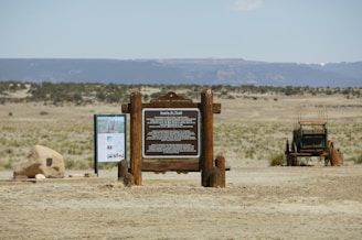 A wooden informational sign stands prominently in a vast desert landscape. Nearby, there is a historical covered wagon, and another informational board displaying maps and text. The background features open plains with sparse vegetation and distant mountains under a clear blue sky.