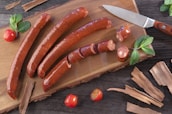 Four smoked sausages rest on a wooden cutting board. One sausage is partially sliced with thick, even cuts. A few sprigs of fresh mint and bright red cherry tomatoes are placed around the board, adding a fresh contrast. A sharp knife with a wooden handle lies beside the sausages. Rustic wooden strips are scattered around, creating a natural and earthy setting.
