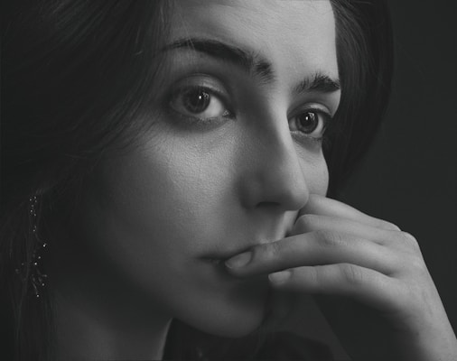 A pensive black-and-white portrait of a woman looking away from the camera. Her fingers gently touch her lips, giving an impression of deep thought or contemplation. The lighting highlights the texture of her skin and the emotion in her eyes.