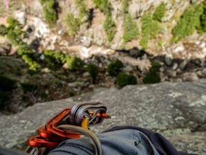 The image captures a view from a high, rocky ledge, looking down into a forested valley. Carabiners and climbing gear are visible in the foreground, suggesting a rock climbing activity. The background shows a mix of green trees and rocky terrain, with a steep drop below.