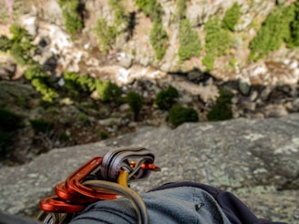 The image captures a view from a high, rocky ledge, looking down into a forested valley. Carabiners and climbing gear are visible in the foreground, suggesting a rock climbing activity. The background shows a mix of green trees and rocky terrain, with a steep drop below.