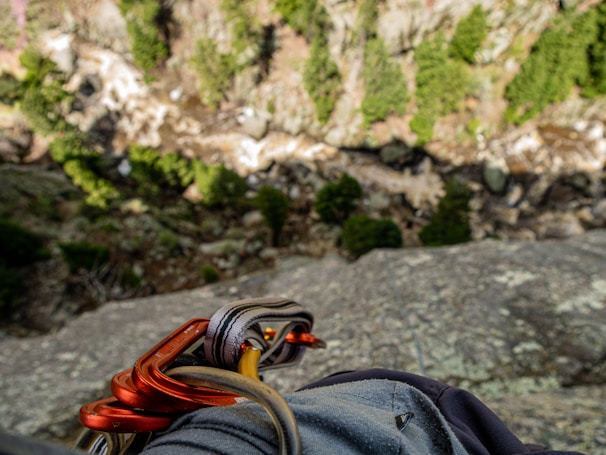 The image captures a view from a high, rocky ledge, looking down into a forested valley. Carabiners and climbing gear are visible in the foreground, suggesting a rock climbing activity. The background shows a mix of green trees and rocky terrain, with a steep drop below.