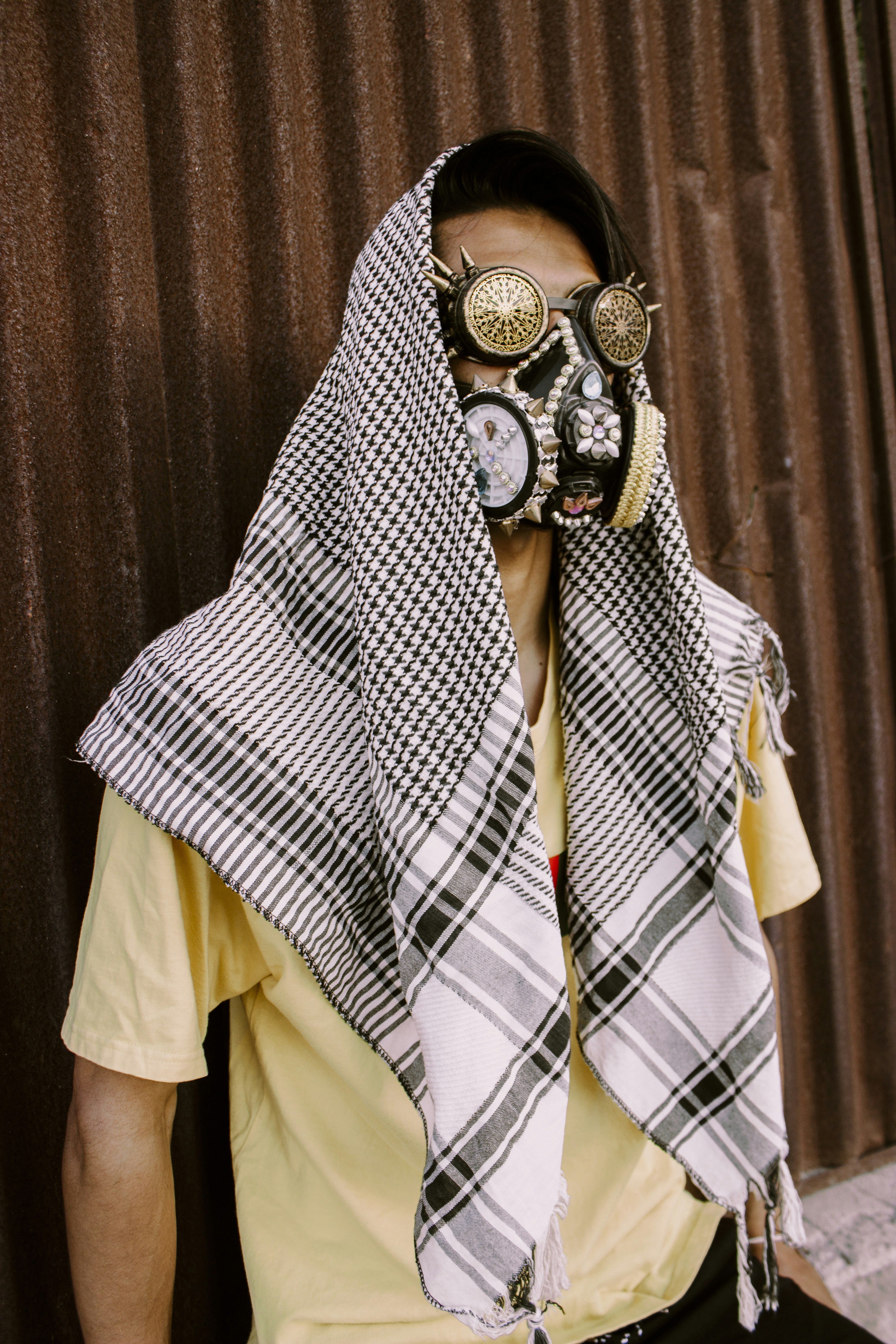 great photo recipe,how to photograph fine art portrait of a male model wearing strange mask and steampunk glasses and a palastenian scarf.; man in blue white and brown plaid button up shirt wearing brown leather face mask
